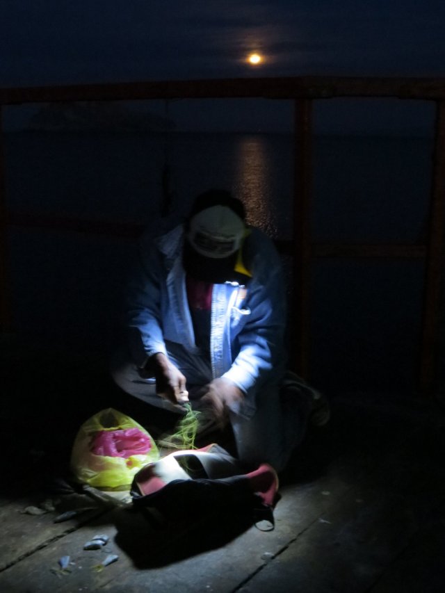 Flashlight tucked under his chin, a fisherman repairs his net.